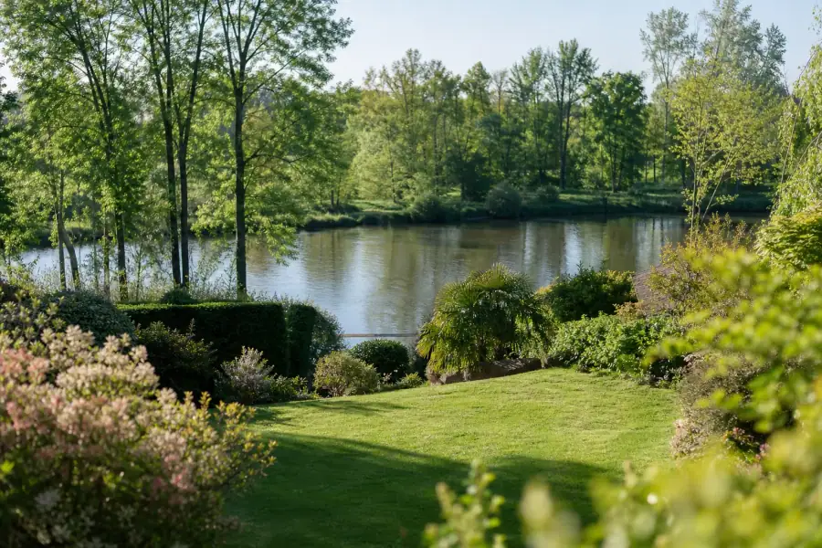 Un jardin pavillonnaire soigné en léger contrebas, pelouse dense d'un vert profond et massifs bien tenus, avec la ligne d'eau calme de la Sarthe bordée d'arbres en arrière-plan. Lumière douce de fin de matinée, ambiance fraîche et vivante de bord de rivière. Cadrage large horizontal qui relie le jardin au fil de l'eau, sans personne ni marque visible.